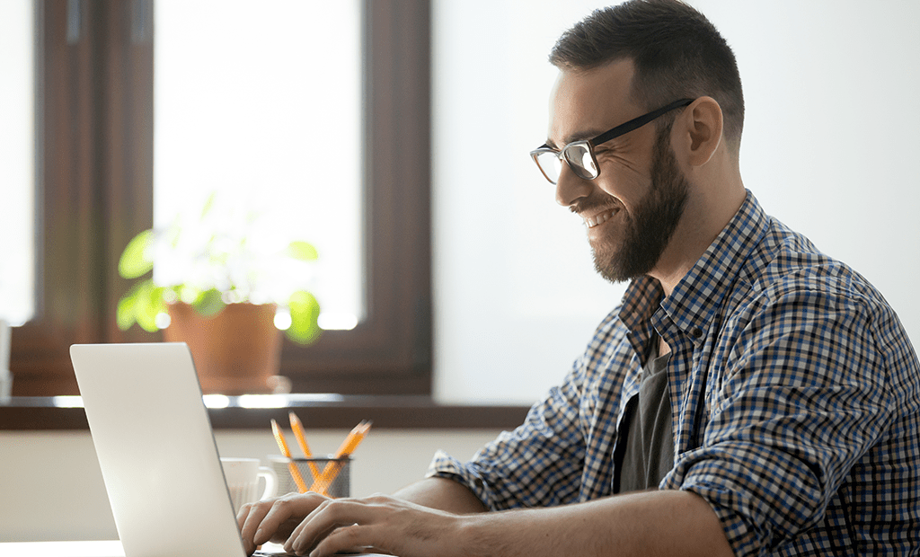 iImpact. Homem jovem, sorrindo enquanto trabalha em um laptop. Ele usa óculos e uma camisa xadrez em tons de azul e branco. No fundo, há uma janela com luz natural entrando, e uma planta em vaso está posicionada no parapeito. 