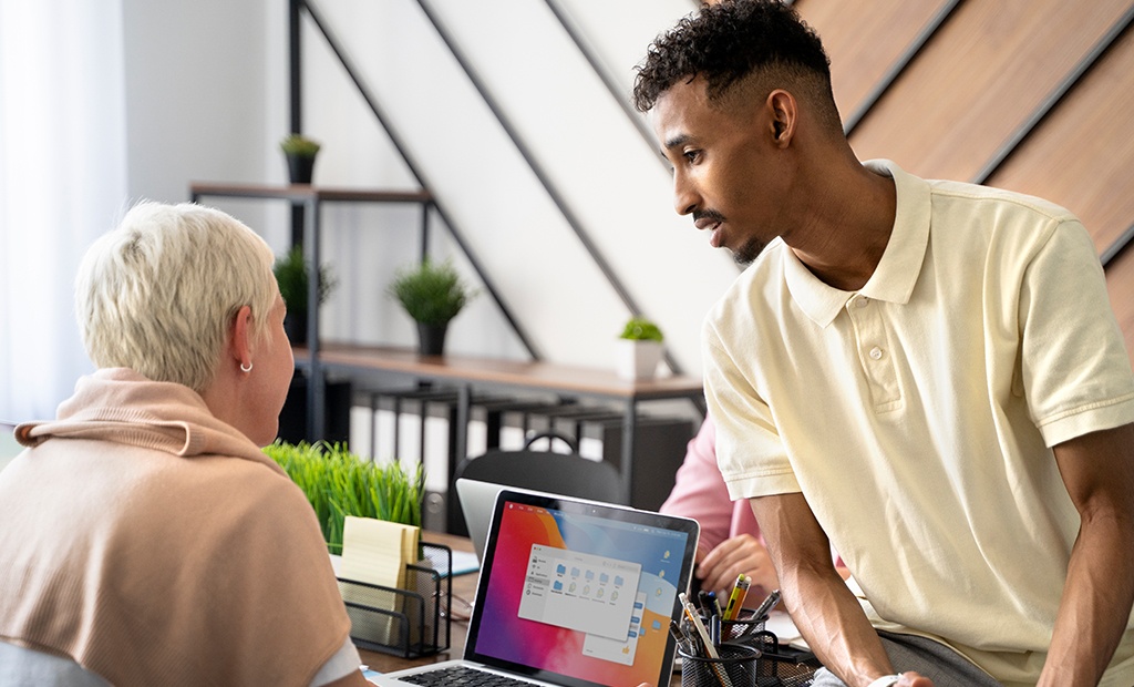 Inclusão digital. Homem de camisa polo clara inclinado em direção a uma mulher de cabelo curto e loiro, que está sentada à mesa. Há um laptop aberto à frente dela.