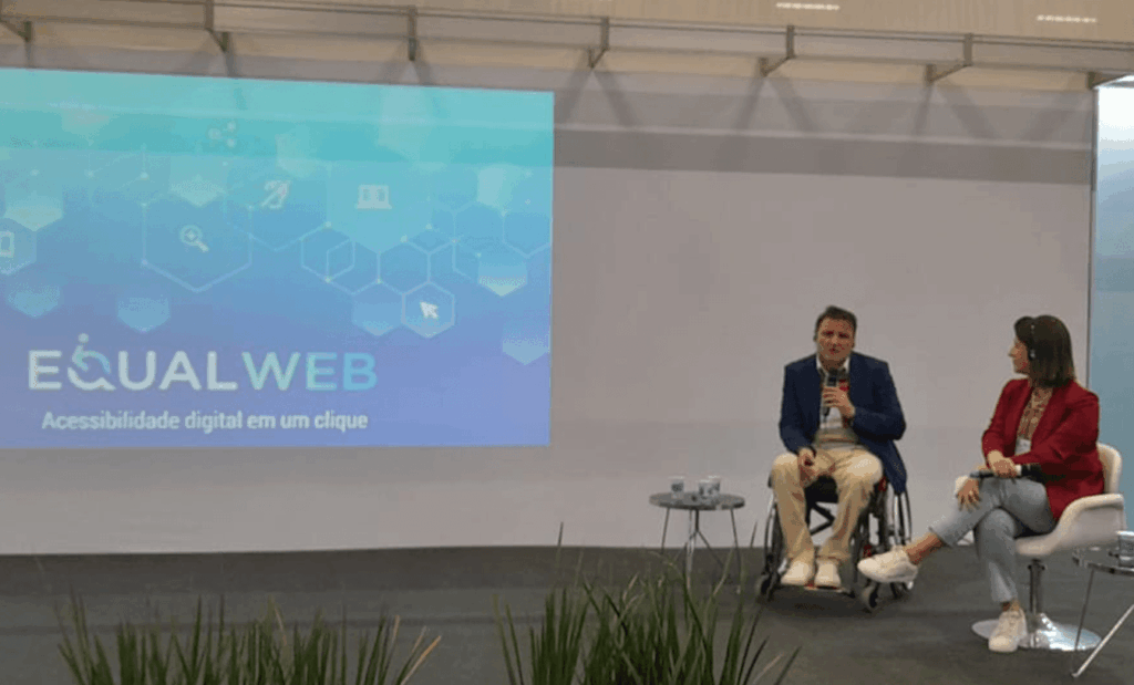 Reatech. No palco temos o Fabio Uzunof, homem branco, de cabelos castanhos e barba grisalha, vestindo calça bege, camisa branca e blazer azul, sentado em sua cadeira de rodas com um microfone na mão. Ao lado dele, temos a Diretora de Operações Marketplace na Via Varejo, Fernanda Winck, mulher branca, de cabelos castanhos em tamanho curto, vestindo calça jeans, camisa bege e casaco vermelho, sentada em uma cadeira branca. Ambos estão sorridentes olhando para a foto.