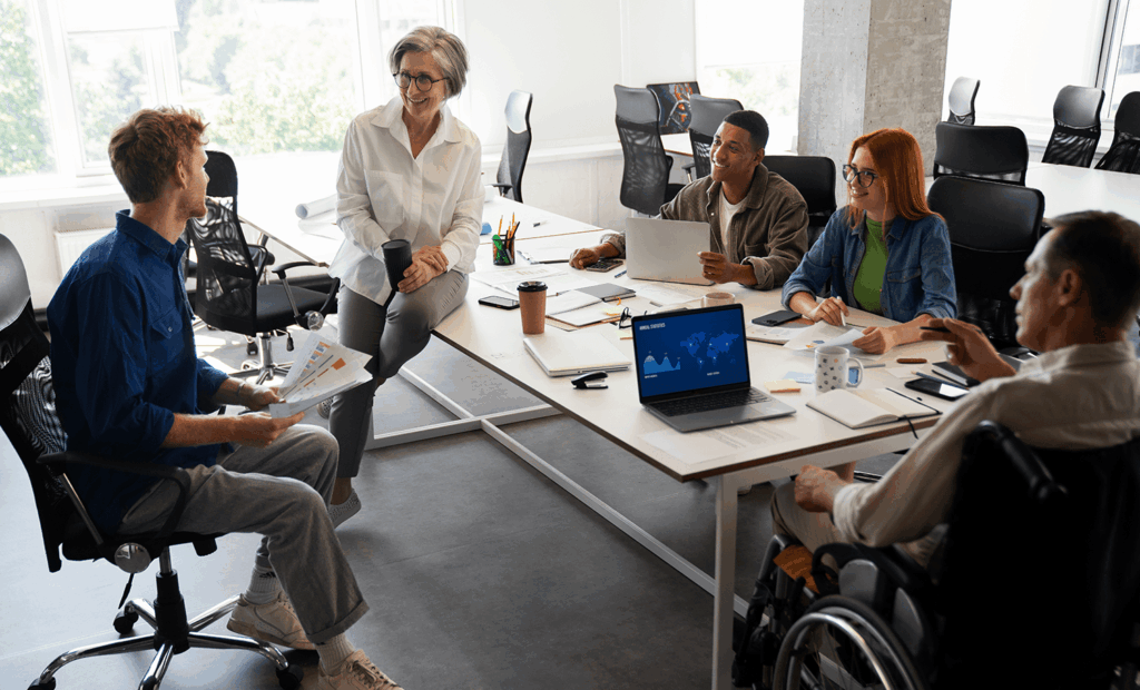 Diversidade. A imagem representa uma sala de reunião com os seguintes integrantes conversando: Um home ruivo sentado em uma cadeira segunrando alguns papeis, uma mulher que aparenta ter uns 60 anos segurando um copo e encoonstada na mesa, um homem negro segurando alguns papeos na mão, uma menina ruiva segurando uma papel e uma caneta e um homem cadeirantes gesticulando com as mãos. 