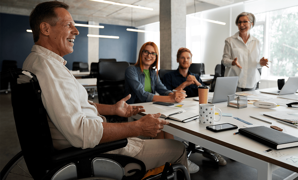 acessibilidade digital. Imagem de uma sala de reunião com pessoas conversando. Na ponta da mesa temos um homem cadeirante, pardo e cabelos grisalhos sorrindo ao seu lado temos uma menina jovem, ruiva e de óculos ao lado remos um menino ruivo, sentado e com olhar pensativo, em pé ao lado temos uma mulher de cabelos grisalhos, blusa branca, sorrindo. 