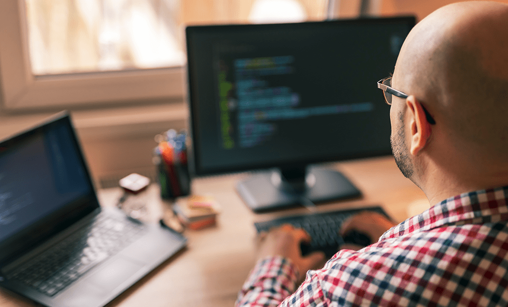 IA. Imagem de um homem branco, careca usando óculos e blusa xadres. Ele esta mexendo no computador e na imagem aparece duas telas a sua frente.