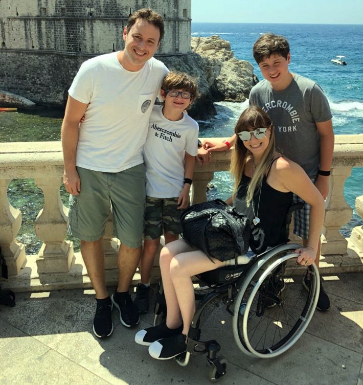 Foto de Jaques Haber e Andrea Schwarz com os dois filhos do casal durante viagem. Ele está em pé, abraçado ao filho menor. Ela está sentada na cadeira de rodas, com o filho maior atrás. A famílias está encostada em uma mureta. ao fundo, o mar e uma construção em pedra.