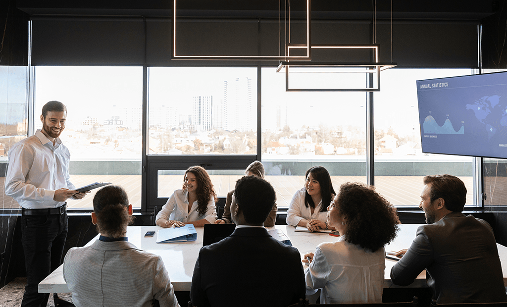 ABNT. Homem de camisa branca em pé, fazendo uma apresentação para pessoas reunidas ao redor de uma mesa em uma sala de reunião.