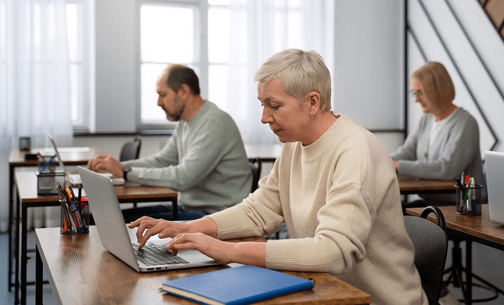Leitores de tela. Três pessoas idosas estão sentadas em mesas individuais em uma sala de aula ou treinamento, cada uma usando um laptop. A mulher em primeiro plano, com cabelo grisalho curto, está concentrada no computador. Atrás dela, um homem e outra mulher também estão trabalhando.